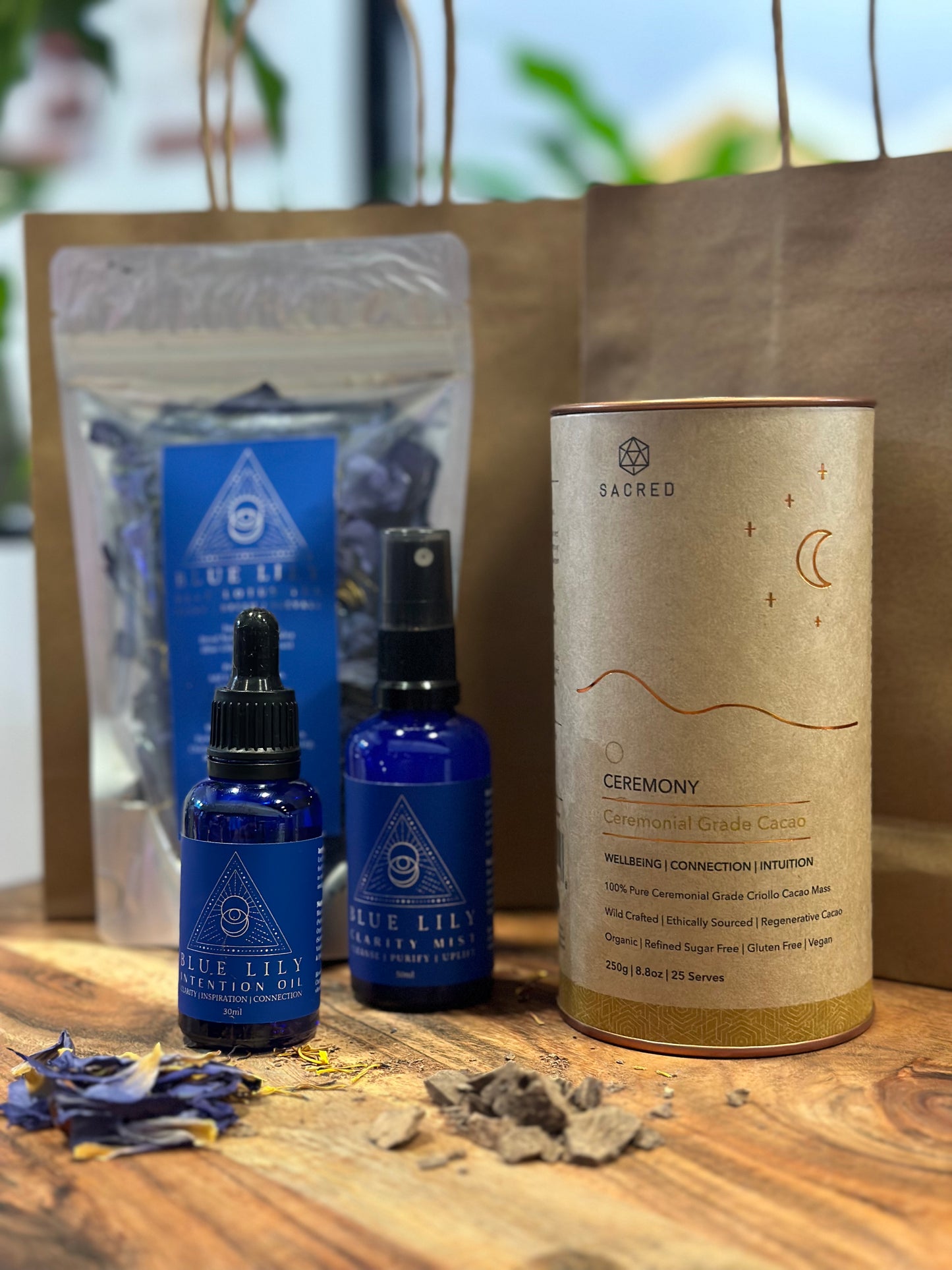 A set of cacao drinking chocolate and blue lotus flower, blue lotus oil, and blue lotus mist, all displayed on a wooden surface with dried loose blue lotus petals around.
