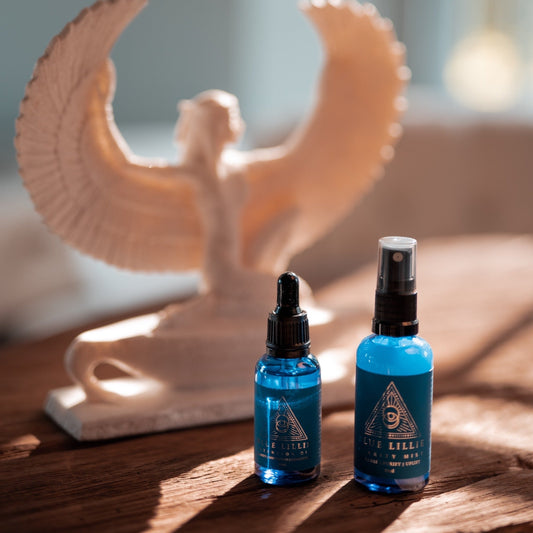 Two blue glass bottles labeled as 'Intention Oil' and 'Clarity Mist' respectively, placed on a wooden surface with a blurred Isis sculpture in the background.