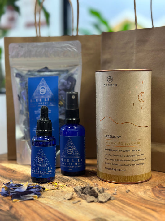 A set of cacao drinking chocolate and blue lotus flower, blue lotus oil, and blue lotus mist, all displayed on a wooden surface with dried loose blue lotus petals around.